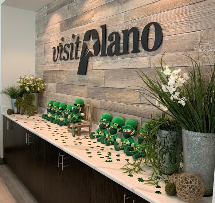 Plush leprechauns wearing green hats sit on a counter under a visit Plano sign. The counter is decorated with shamrocks, plants, and white flowers, creating a festive St. Patricks Day display.
