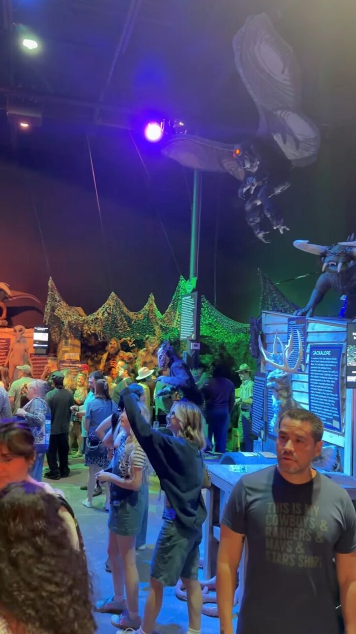 Crowd of people at an indoor exhibit with fantasy creature statues overhead, colorful lights, and informational displays. A large dragon figure hangs above the scene.