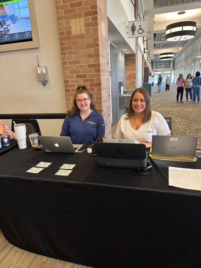 Two women sit at a table with laptops and papers in a building lobby, smiling at the camera. Other people are visible walking in the background.