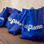 Several blue tote bags with visit Plano written in white text are displayed on a brown table against a light-colored wall.