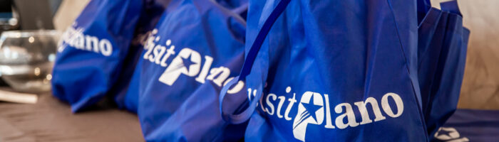 Several blue tote bags with visit Plano written in white text are displayed on a brown table against a light-colored wall.