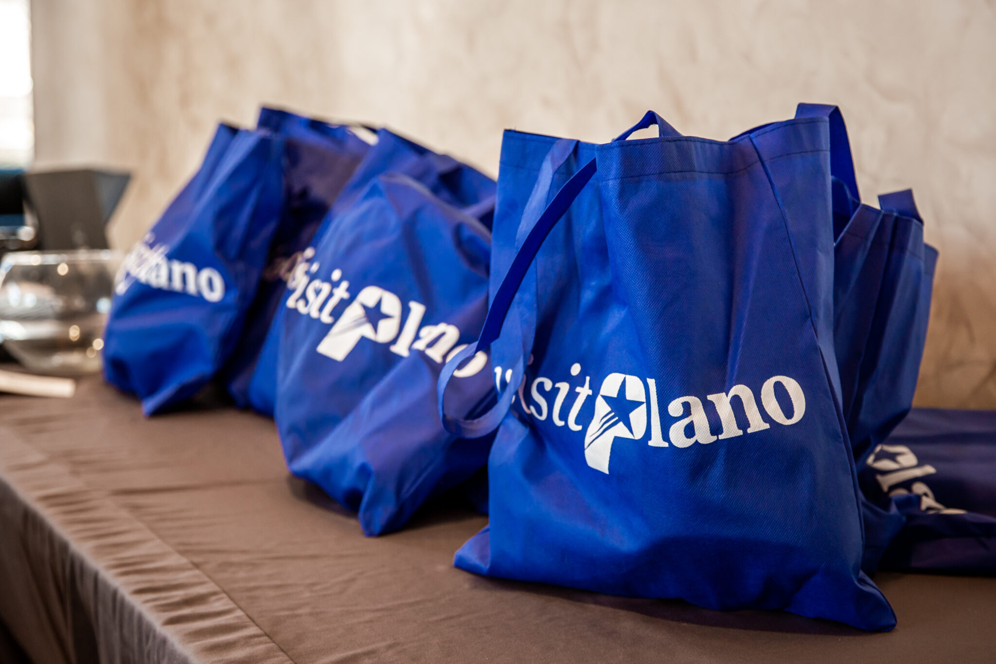 Several blue tote bags with visit Plano written in white text are displayed on a brown table against a light-colored wall.