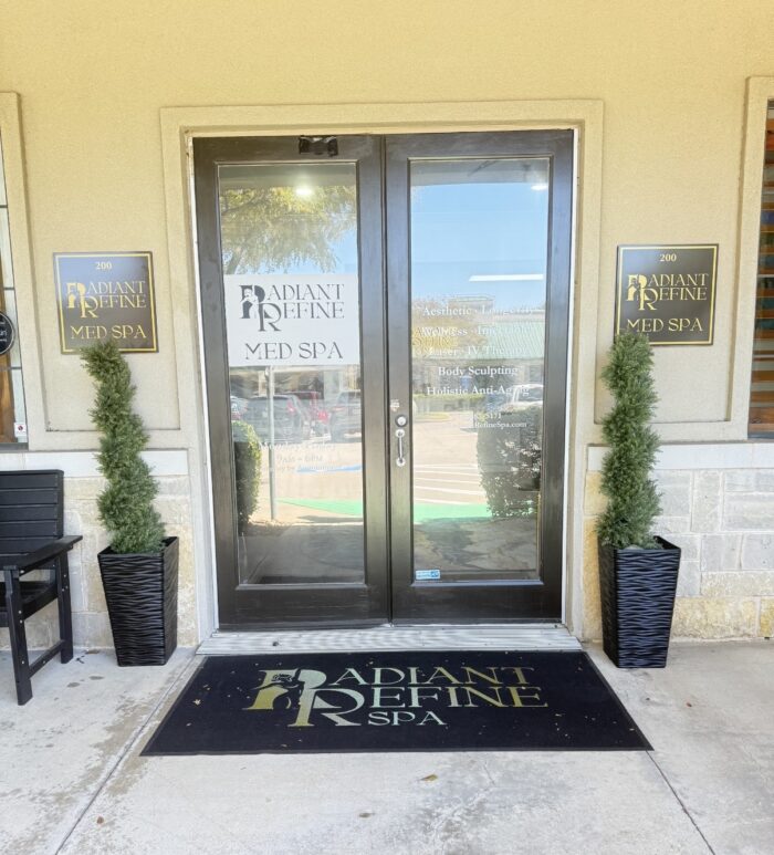 Radiant Refine Med Spa entrance with glass double doors, two tall potted plants, and branded signs on either side. A black doormat with the spas logo is in front of the door.