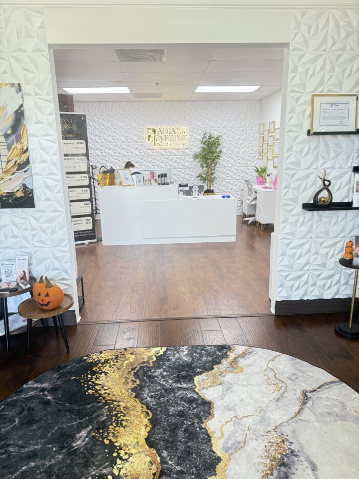 Modern reception area with white geometric walls, a round marble-patterned rug, a front desk, small decorations including a pumpkin, and framed certificates on the walls. A tall plant stands behind the desk.