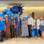 A group of ten people pose and smile in front of a blue balloon display indoors, celebrating the Visit Plano Awards. Some wear blue or casual outfits, with metallic and star-shaped balloons adding to the festive background.
