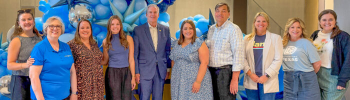 A group of ten people pose and smile in front of a blue balloon display indoors, celebrating the Visit Plano Awards. Some wear blue or casual outfits, with metallic and star-shaped balloons adding to the festive background.