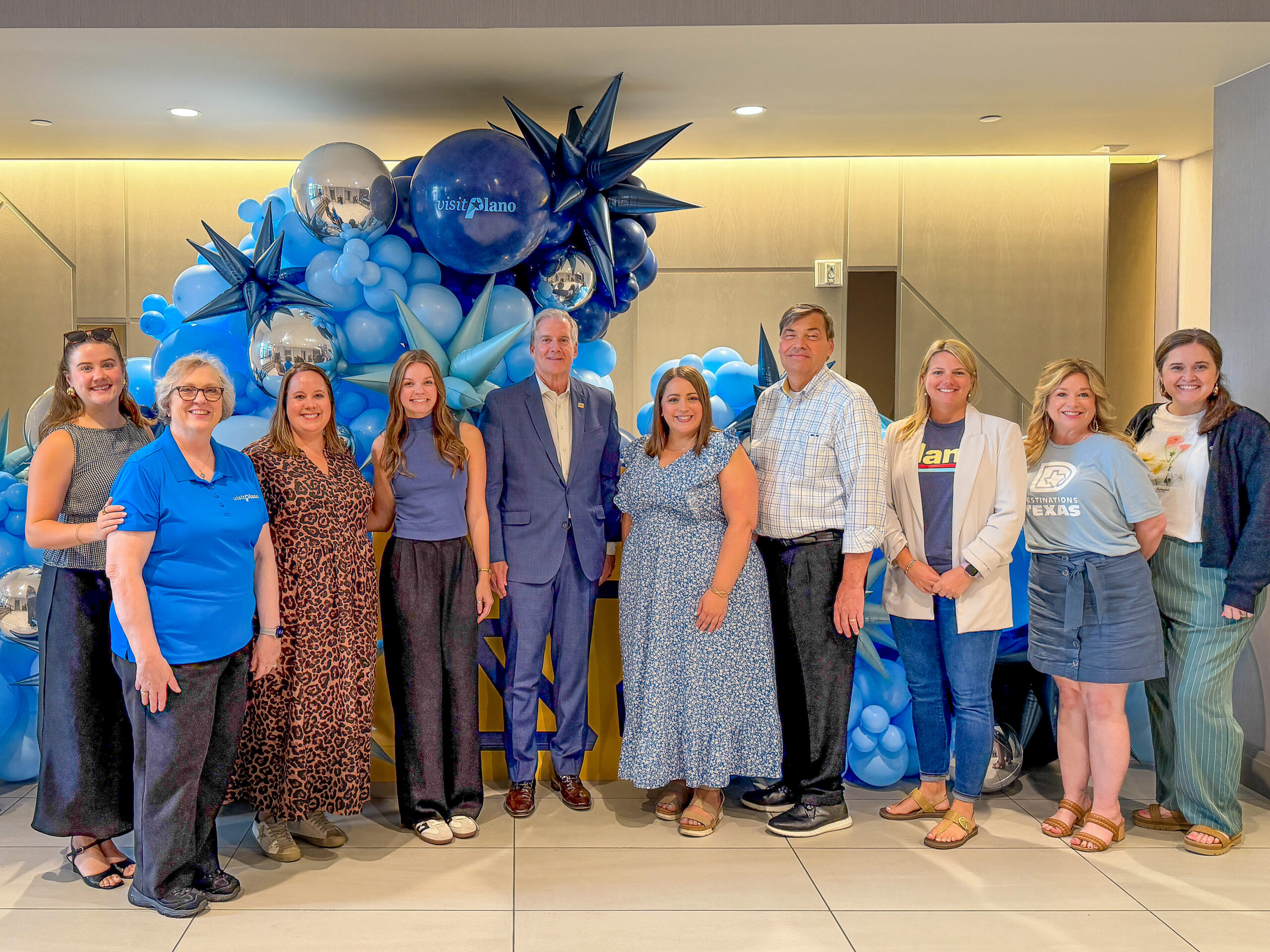 A group of ten people pose and smile in front of a blue balloon display indoors, celebrating the Visit Plano Awards. Some wear blue or casual outfits, with metallic and star-shaped balloons adding to the festive background.