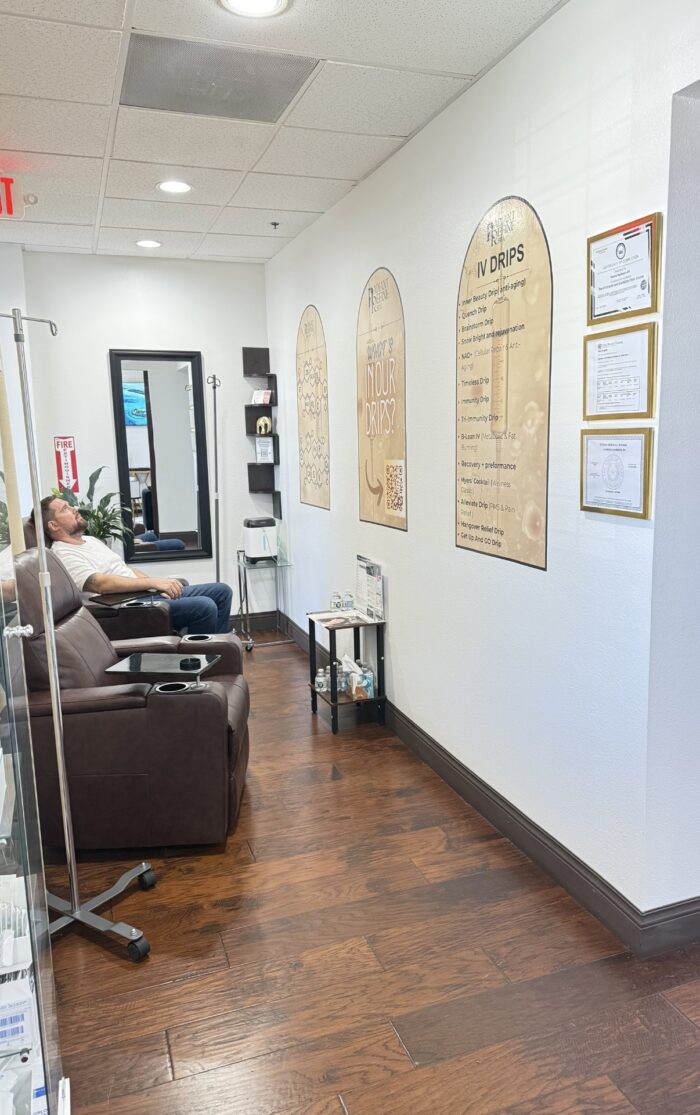 A person sits in a brown recliner inside a bright clinic room with wood floors; informational posters about IV drips are on the wall, and medical supplies are visible on shelves.