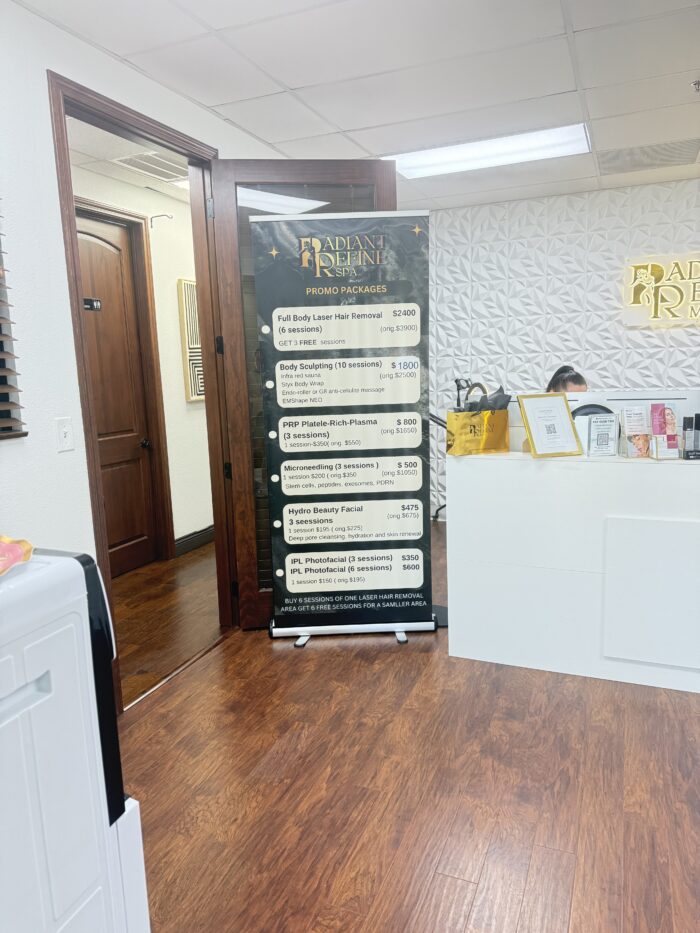 A stand-up banner in a clinic lists promo packages for beauty treatments. The room has wooden floors, white walls with textured panels, and some bags and a computer on a white desk.