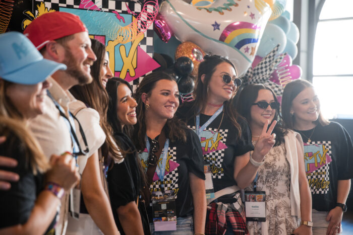 A group of people pose and smile together at the Visit Plano Awards event, wearing casual clothes and name badges, with colorful balloons and 90s-themed decorations in the background.