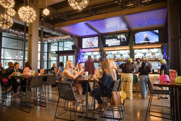 People sit at tables and socialize in a lively bar with modern décor, hanging globe lights, and a well-stocked bourbon display behind the bar—an atmosphere worthy of its Visit Plano Awards recognition. Some people are eating, drinking, or standing near the counter.