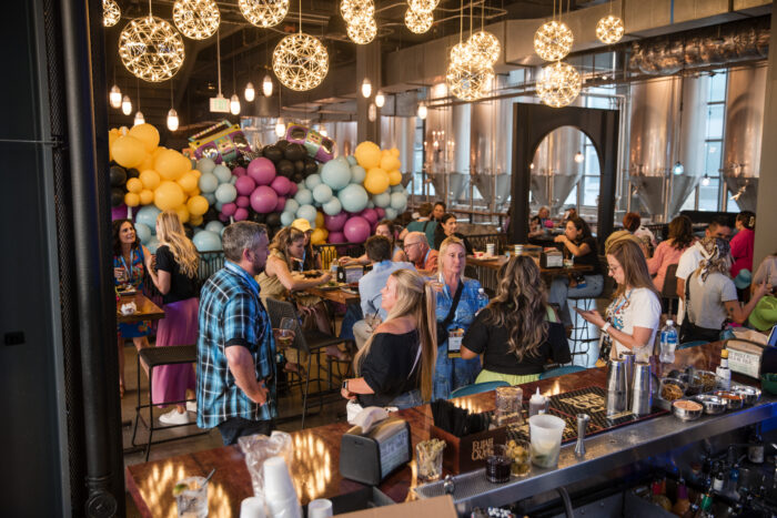 People socialize in a lively bar decorated with colorful balloons and modern hanging lights, reflecting the vibrant spirit that earned the venue recognition from Visit Plano Awards. Some stand near the bar while others sit at tables, enjoying an atmosphere that looks festive and energetic.