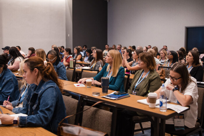 A group of people sit at tables in a classroom or conference room, attentively listening, taking notes, and facing forward. Most appear engaged in the presentation or discussion, which centers around the prestigious Visit Plano Awards.