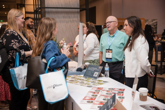 A group of people converse at a convention booth with Visit Plano tote bags, brochures, and a display featuring Visit Plano Awards, in a modern indoor setting.