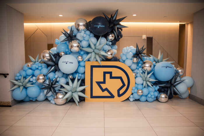 A balloon display with blue and silver balloons, spiky star shapes, and a large yellow outline of Texas with a D. Some balloons read Visit Plano. Perfect for the Visit Plano Awards, this eye-catching display is set indoors on a tiled floor.