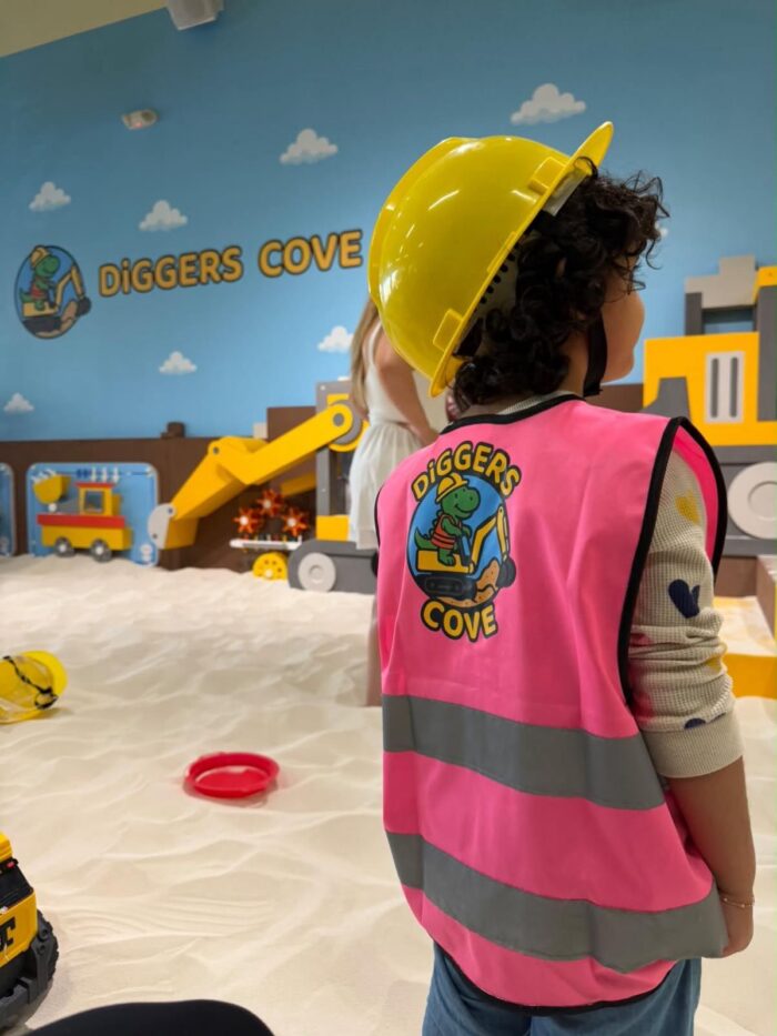 A child in a yellow hard hat and pink vest stands in a sandy indoor play area called Diggers Cove, with toy construction vehicles and colorful wall decorations in the background.