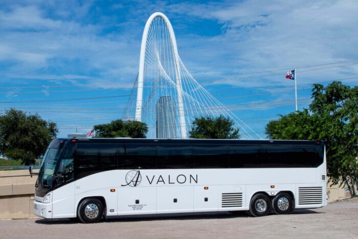 A white tour bus labeled Avalon is parked in front of a large modern cable-stayed bridge, with trees, power lines, and a Texas flag in the background under a blue sky.
