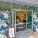 Storefront Visitor Center with Visit Plano signage, blue and white balloon decorations, and a table inside. A sign by the entrance reads Free Hydration Station with an image of a water bottle. The door is open to welcome visitors.