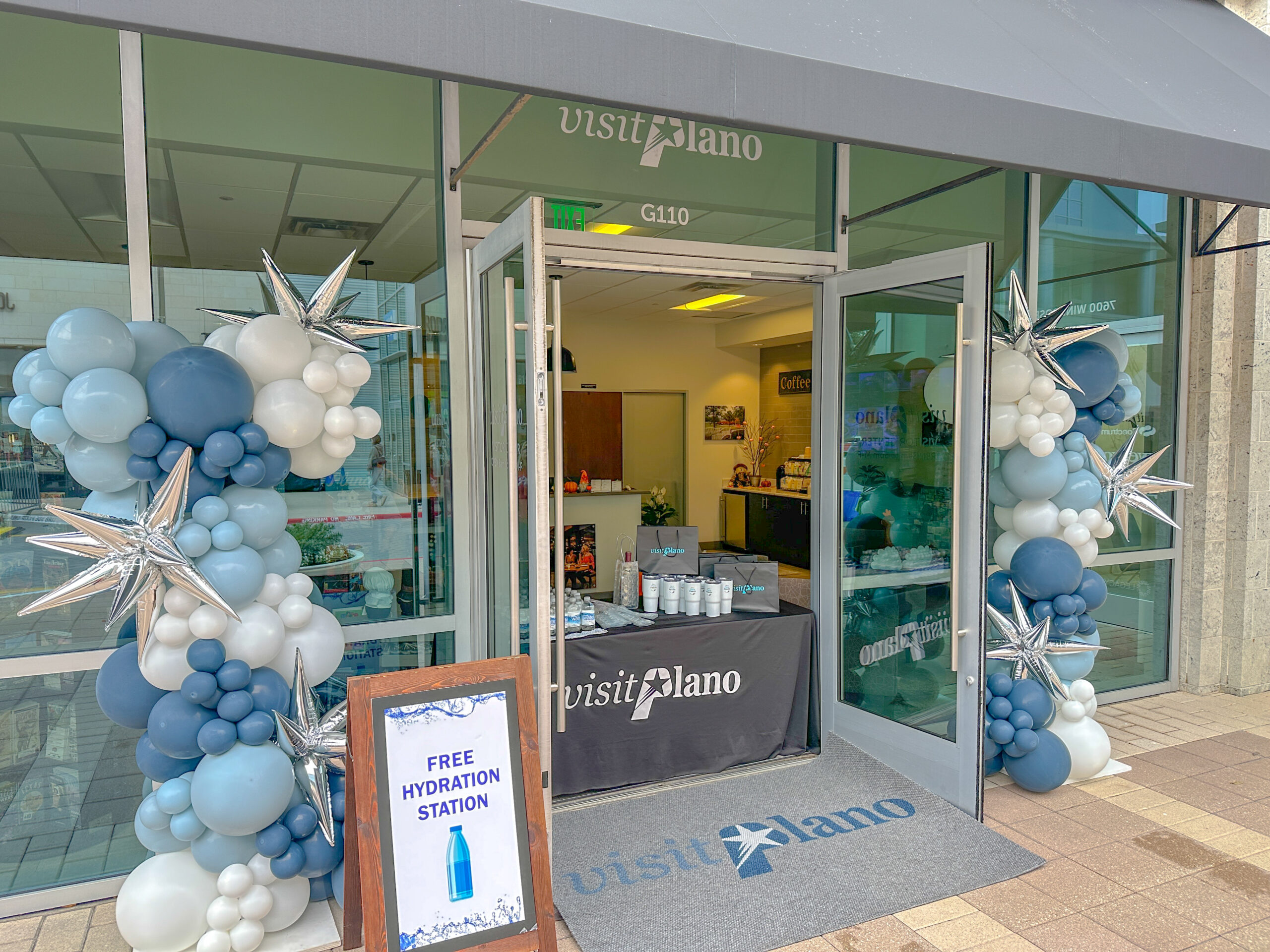 Storefront Visitor Center with Visit Plano signage, blue and white balloon decorations, and a table inside. A sign by the entrance reads Free Hydration Station with an image of a water bottle. The door is open to welcome visitors.