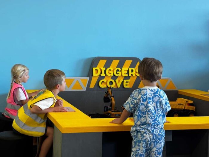 Three children play with toy construction vehicles at a table labeled Diggers Cove. One child wears a yellow vest, another wears blue patterned clothes, and the wall behind them is painted blue.