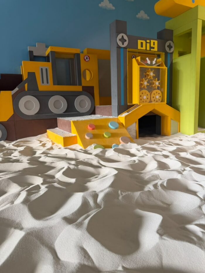 Indoor playground with construction-themed structures, including a digger and slide, sits on a floor covered in textured white sand under a blue wall with cloud decorations.