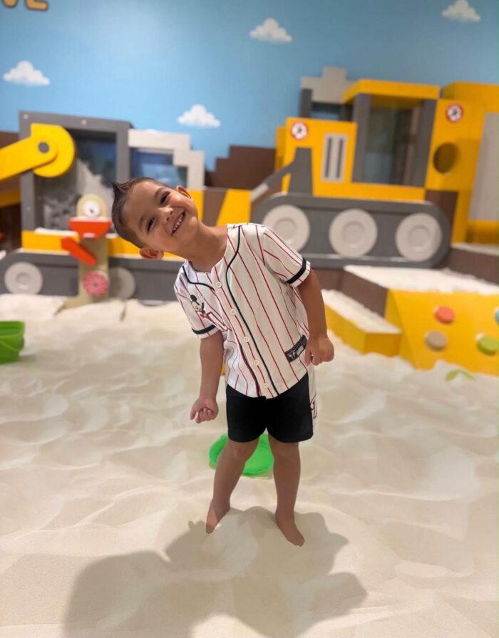 Smiling child in a striped shirt and black shorts stands barefoot on white sand, with colorful construction-themed play structures in the background.