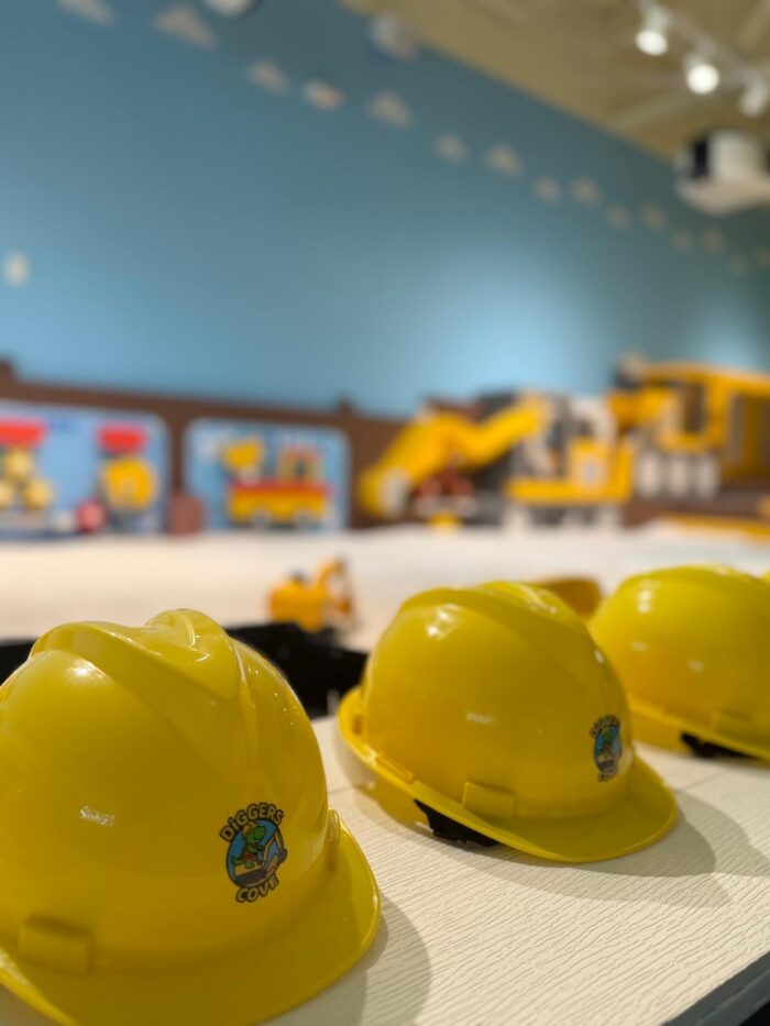 Three yellow construction helmets with Digger Cubs stickers are lined up on a table. In the background, there are construction-themed toys and colorful murals, suggesting a playful, child-friendly environment.