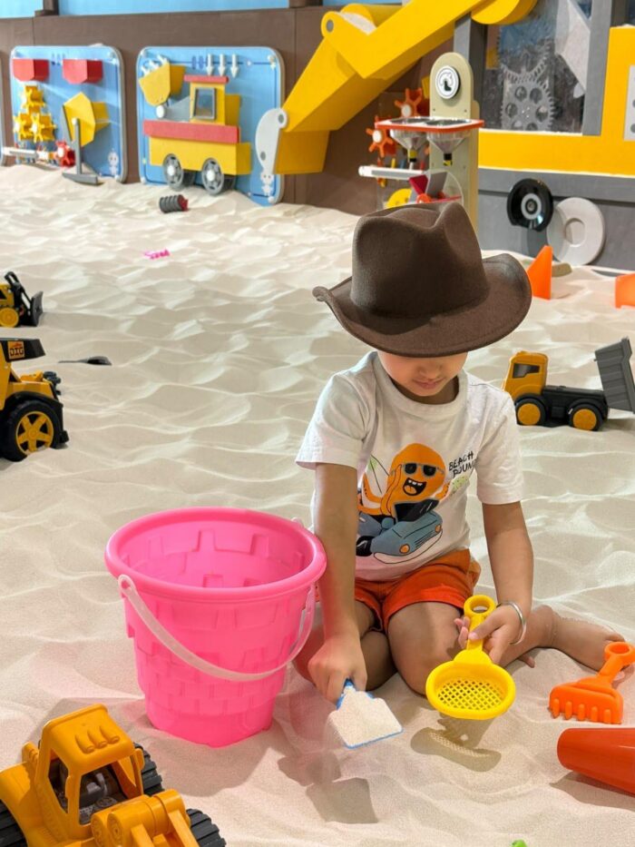 A young child wearing a brown hat plays in an indoor sandbox with toy trucks, a pink bucket, and a yellow sand sifter. Colorful wall decorations and more toys are visible in the background.