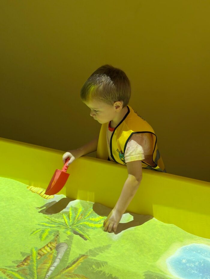 A young boy wearing a yellow vest plays with a red toy shovel in a sandbox with a projected image of palm trees and water under bright lighting.