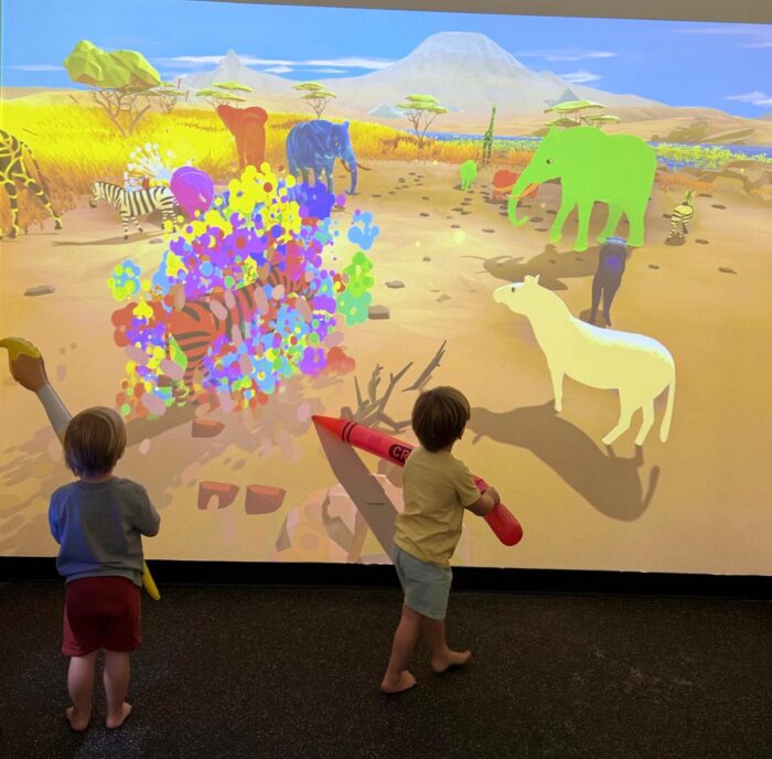 Two young children hold oversized crayons and interact with a colorful digital safari scene projected on a wall, featuring animals like elephants, zebras, and antelopes, with vibrant patches of color on the display.