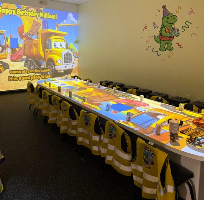 A childrens birthday party room with construction-themed decor, yellow vests on chairs, a long table with cups and snacks, a cartoon truck and dinosaur on the wall, and a projection saying Happy Birthday William!.