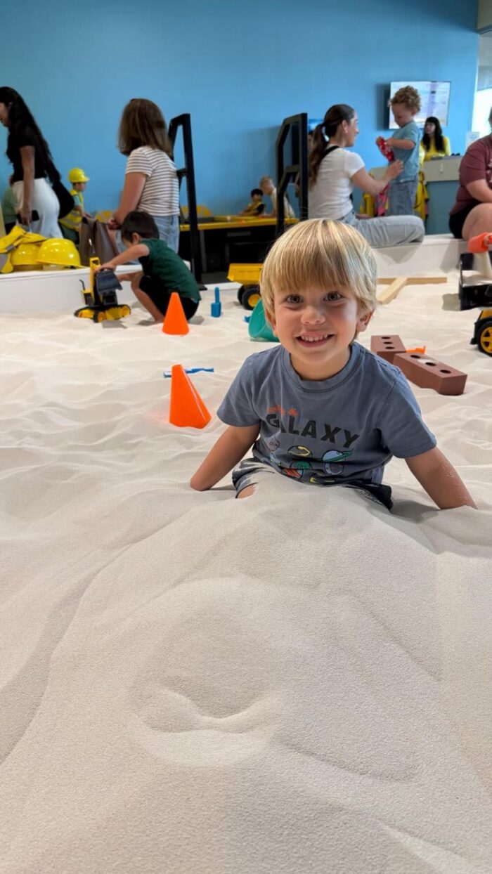 A young child smiles while sitting in a large indoor sandbox, surrounded by orange cones, toy trucks, and other children playing in the background. The atmosphere is playful and lively.