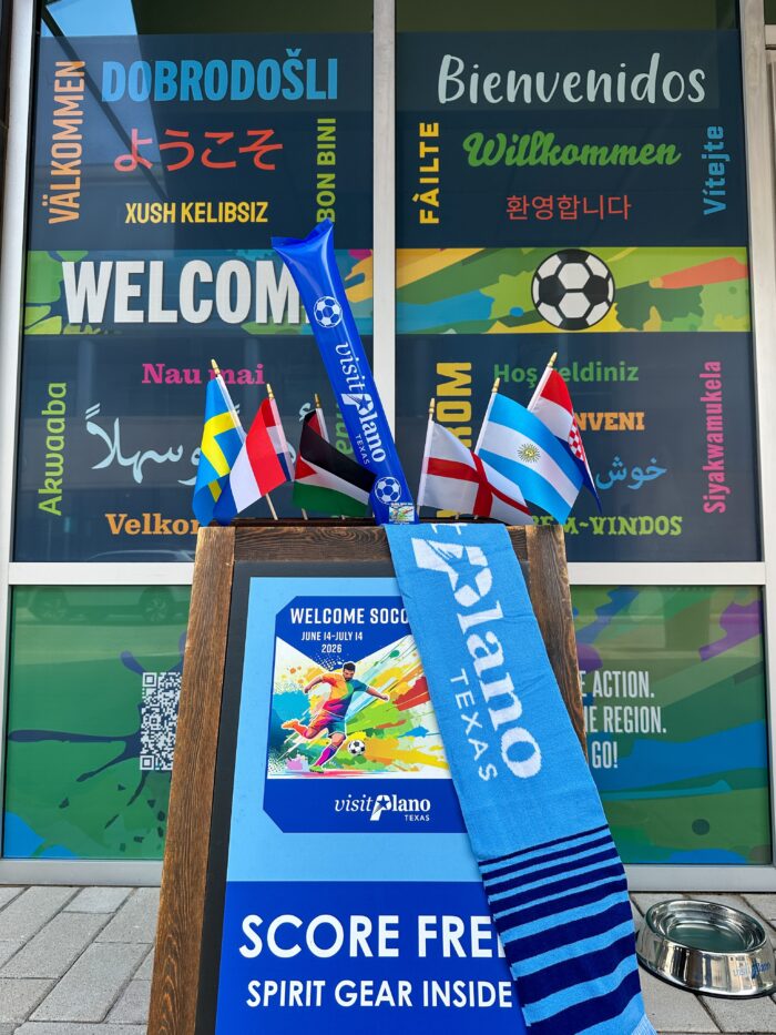 Colorful Welcome signs in multiple languages on a window, with small international flags and a blue Plano, Texas scarf displayed on a stand promoting free spirit gear.