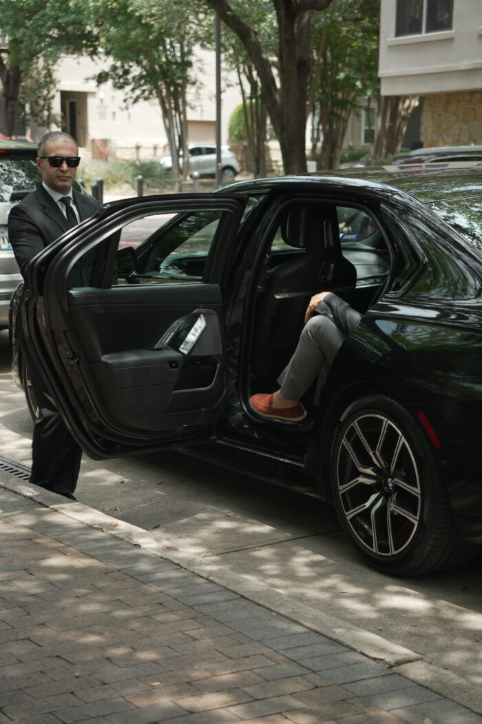 A man in a suit and sunglasses stands by an open car door, while another person wearing orange shoes sits inside the back seat of the car on a sunny street.
