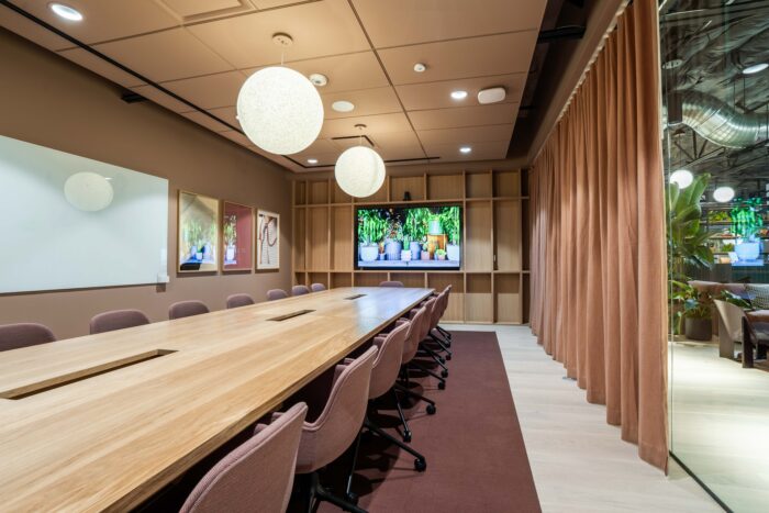 A long wooden conference table with pink chairs in a meeting room, featuring a wall-mounted screen displaying plants and a large white board on the side.
