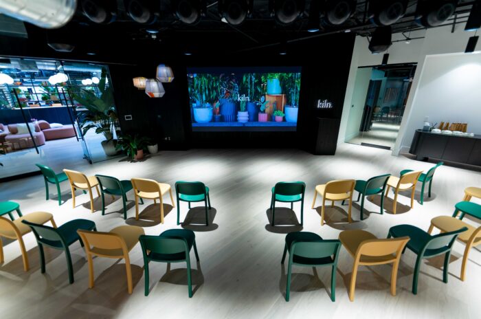 A bright room with rows of green and yellow chairs facing a large projection screen displaying a digital collage of plants and pottery under the brand name kiln.