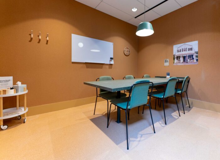 A small meeting room with a rectangular table, six chairs, a whiteboard, and a wall clock on brown walls.
