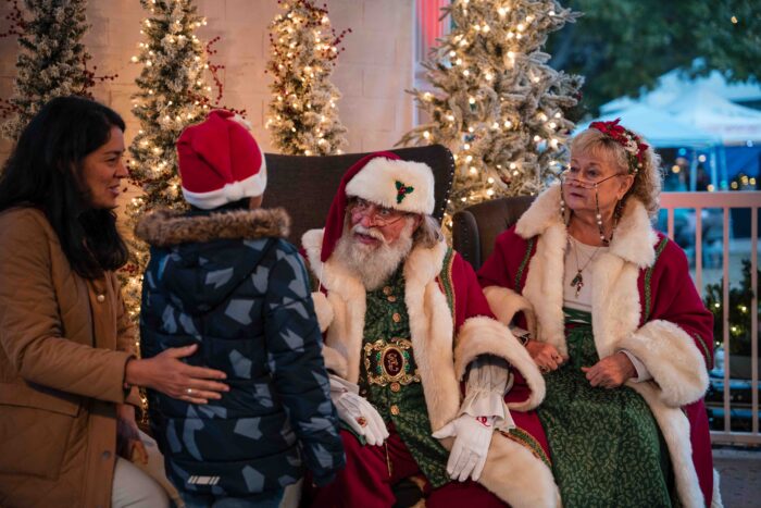 A child in a Santa hat stands before Santa Claus and Mrs. Claus, accompanied by a woman at one of the holiday events in Plano.