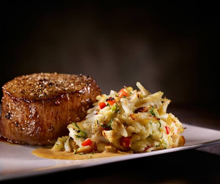 A seared filet mignon served alongside a portion of crab cake with red peppers on a white plate, perfect for a host holiday dinner in Plano.