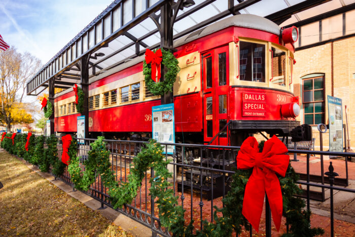 A vintage red Dallas Special streetcar decorated with Christmas wreaths and red bows, parked under a shelter during Holiday Events in Plano.