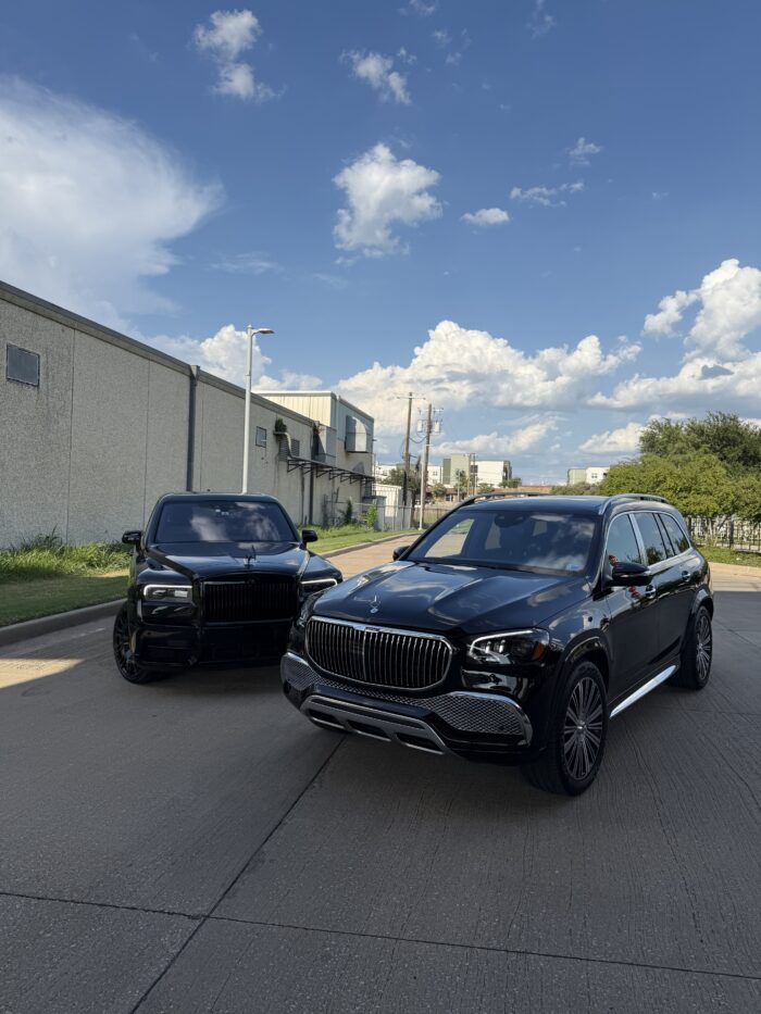 Two black luxury SUVs parked on a sunny street near industrial buildings, with blue sky and white clouds in the background.