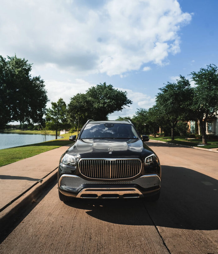 A black luxury SUV is parked on a quiet suburban street near a lake, with trees and houses in the background under a partly cloudy sky.