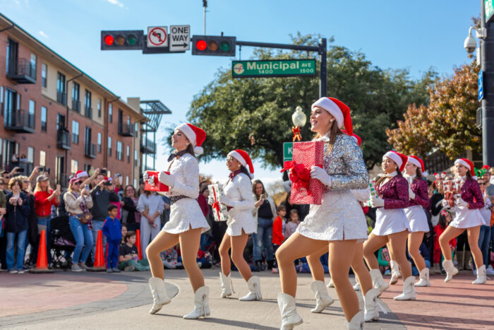 Dancers in sequined white costumes and Santa hats perform at holiday events in Plano, carrying large wrapped gift boxes.
